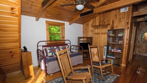 A cozy cabin room with a wooden bed, a rocking chair, a bookshelf, and a window with a view of greenery. - Riverfront New River Cabin - Relax and Renew - Family Cabin Near Boone (Todd)