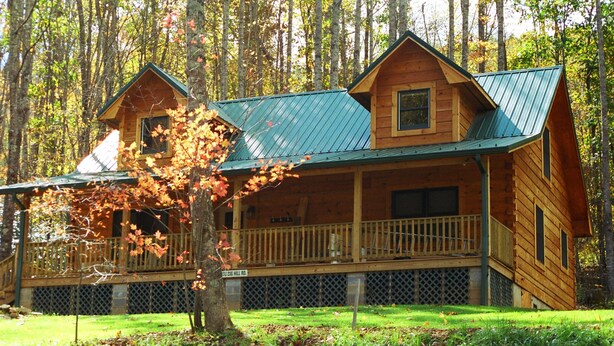 A log cabin with a green metal roof, a porch, and a view of a forested area. - Riverfront New River Cabin - Relax and Renew - Family Cabin Near Boone (Todd)