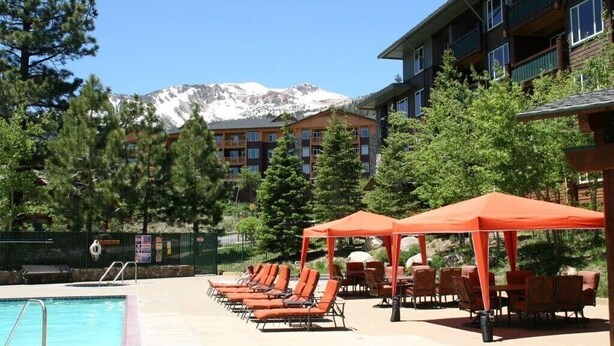 A pool area with lounge chairs and umbrellas, a building with balconies, and a mountain backdrop.
