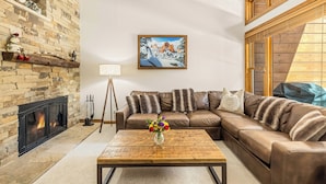 A cozy living room with a stone fireplace, a brown leather sofa, a wooden coffee table with a vase of flowers, and a painting of a snowy mountain scene.