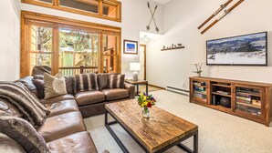 A living room with a sectional sofa, a coffee table with a vase of flowers, a wooden entertainment center, and a large window with blinds.