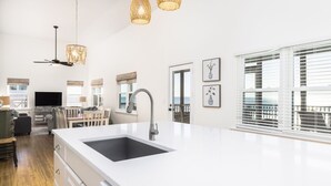 A modern kitchen with a large island, white cabinetry, and a black sink. There are wicker pendant lights, a ceiling fan, and a view of the ocean through large windows. - Stay @ Sea Cookie! Classic, Updated Beach Home (Folly Beach)