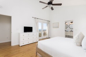 Quarto com cama, televisão sobre uma cômoda branca, ventilador de teto, porta para varanda e banheiro com espelho e pia.