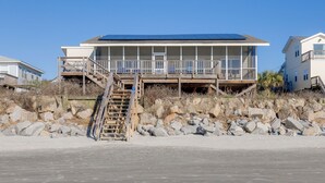 A beachfront house with a wooden staircase leading to a balcony, situated on a rocky shoreline. - Stay @ Sea Cookie! Classic, Updated Beach Home (Folly Beach)