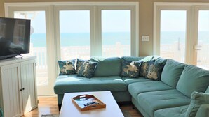 A living room with a teal sofa, a flat-screen TV on a cabinet, and a view of the ocean through large windows.