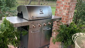 A Char-Briul gas grill with four burners, a side shelf, and a chimney, set on a patio with potted plants and a white metal chair.