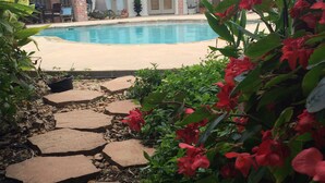 A pool area with a stone pathway leading to a pool, surrounded by greenery and a building with a tiled roof.