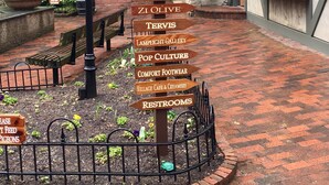 A brick pathway with directional signs pointing to various local attractions and amenities such as The Honey Pot, Life is Good, The Silver Tree, Zi Olive, Tervis, Lamplight Gallery, Pop Culture, Comfort Footwear, Village Cafe & Creamery, and Restrooms.