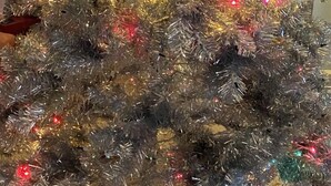 Un sapin de Noël argenté, décoré de guirlandes lumineuses multicolores, se trouve dans une pièce ornée d’un lustre et comprenant une table à manger.