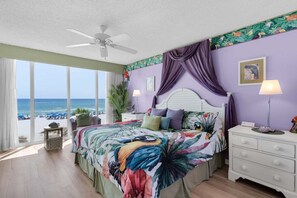 A bedroom with a large bed, a nightstand, a ceiling fan, and a view of the beach.