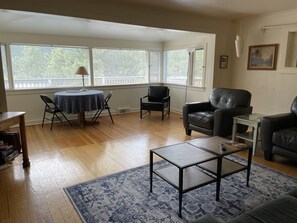 A living room with a wooden floor, a round table with chairs, a sofa, and a coffee table. There are large windows with blinds, a lamp, and a painting on the wall.