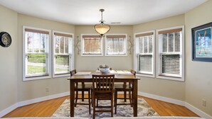 A dining area with a wooden table, four chairs, a chandelier, and large windows with blinds. - Wintergreens R611- Mtn Condo/View, Gated Resort, Indoor/Outdoor Pools & Hot Tub (Banner Elk)