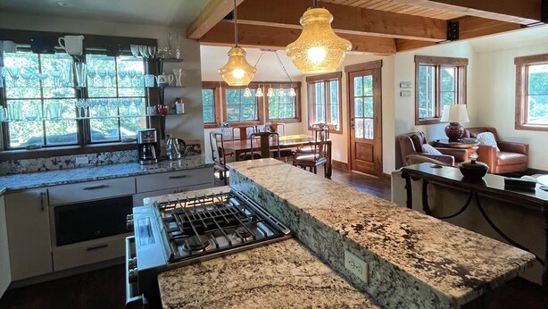 A kitchen with granite countertops, a gas stove, hanging pendant lights, and a dining area with wooden furniture.