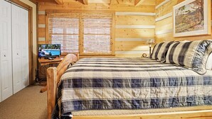 A wooden cabin bedroom with a bed, a desk with a computer, and a window with blinds.