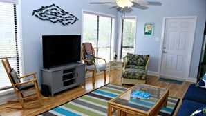 Ein Wohnzimmer mit Flachbildfernseher, zwei Schaukelstühlen, einem Blumenmuster-Sessel, einem Glastisch und einem Streifen Teppich.