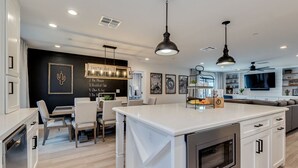 A modern kitchen with a large island, white cabinetry, and stainless steel appliances. There is a dining area with a table and chairs, and a black accent wall with framed artwork.