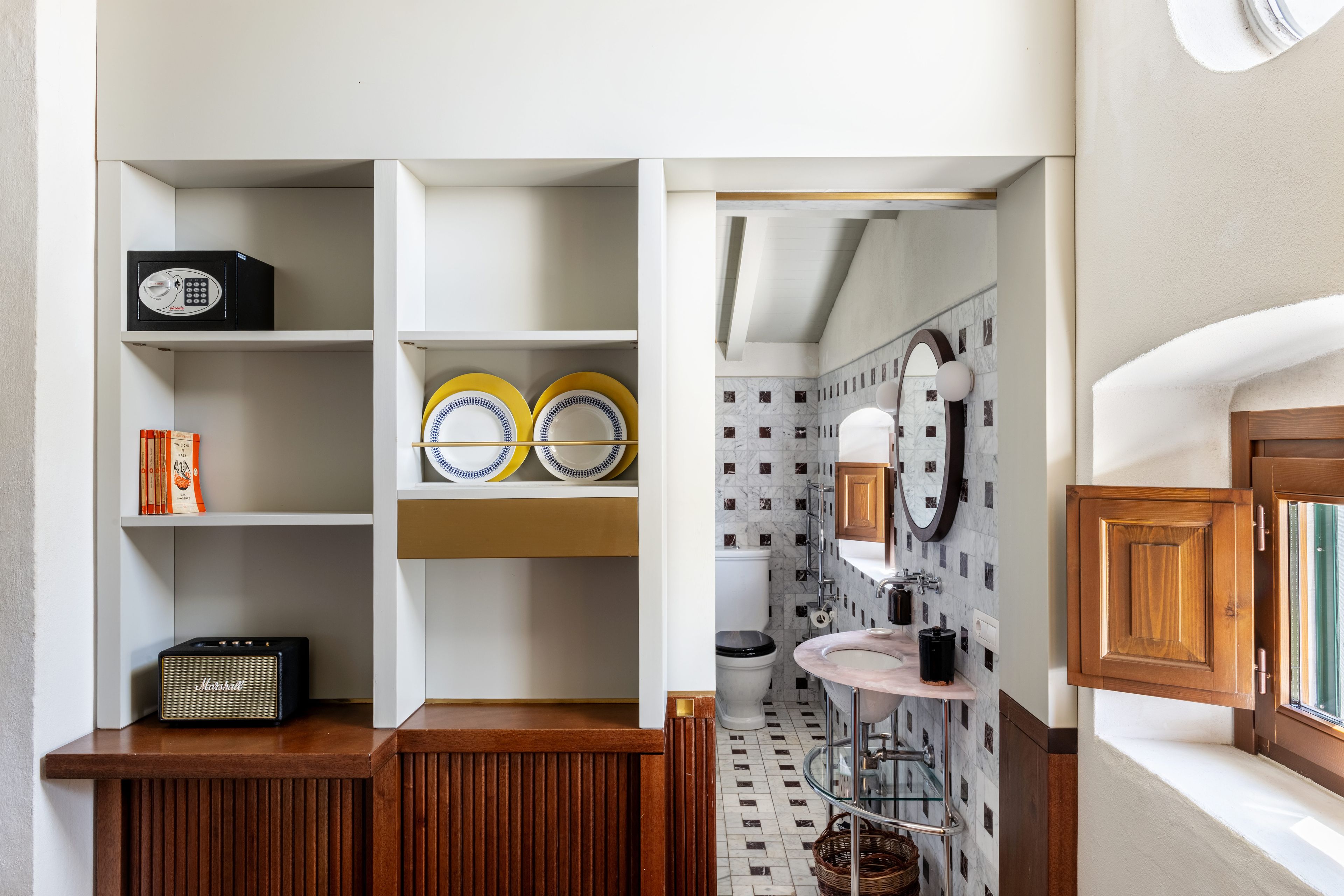 A compact bathroom with a toilet, sink, and mirror. There is a shelf with books and a radio.