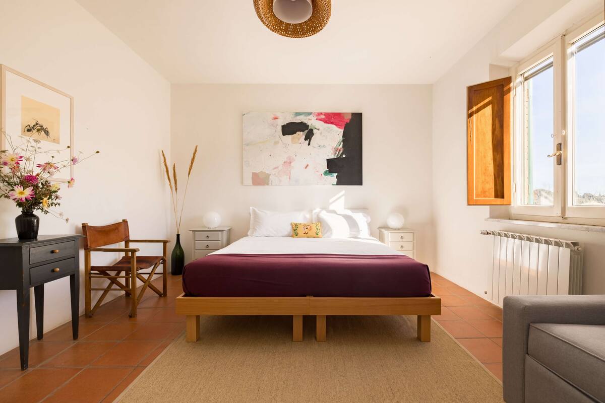 Un dormitorio con cama de madera, un escritorio con silla, un florero con flores y una ventana con vistas.