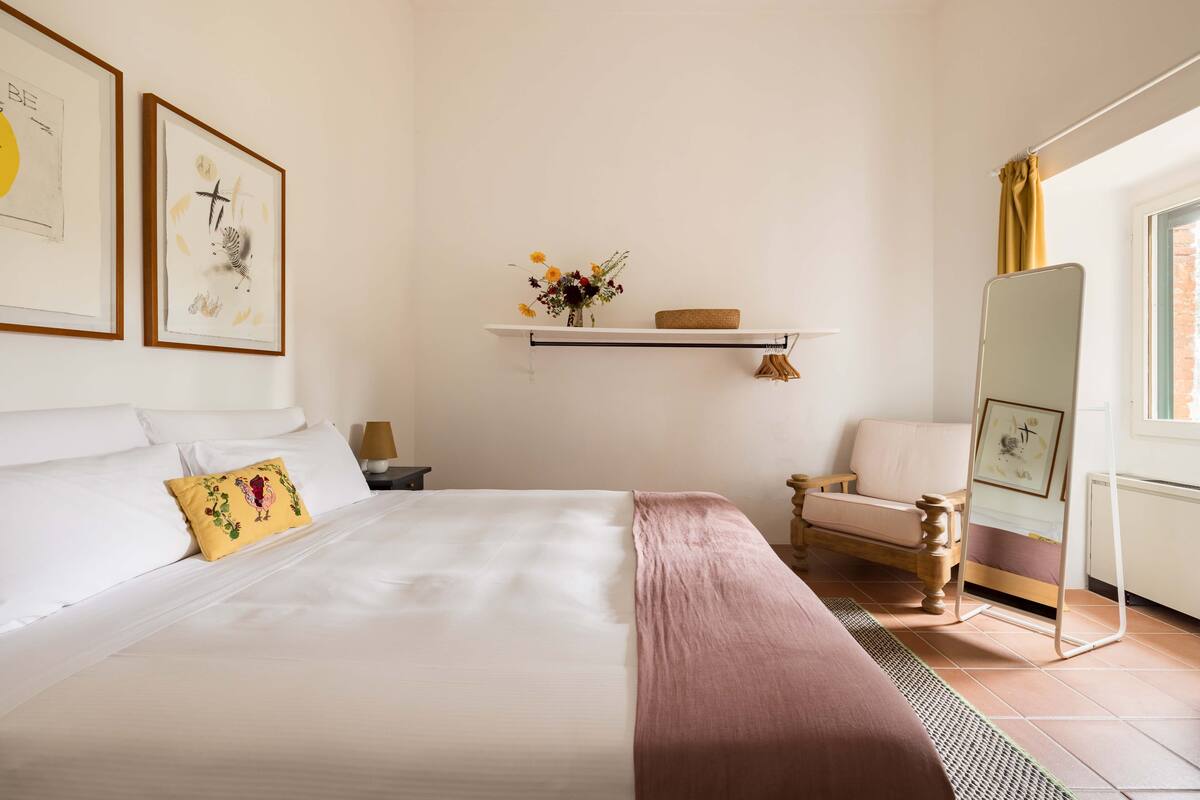 Un dormitorio ordenado con una cama, una silla, una estantería con flores y un espejo.