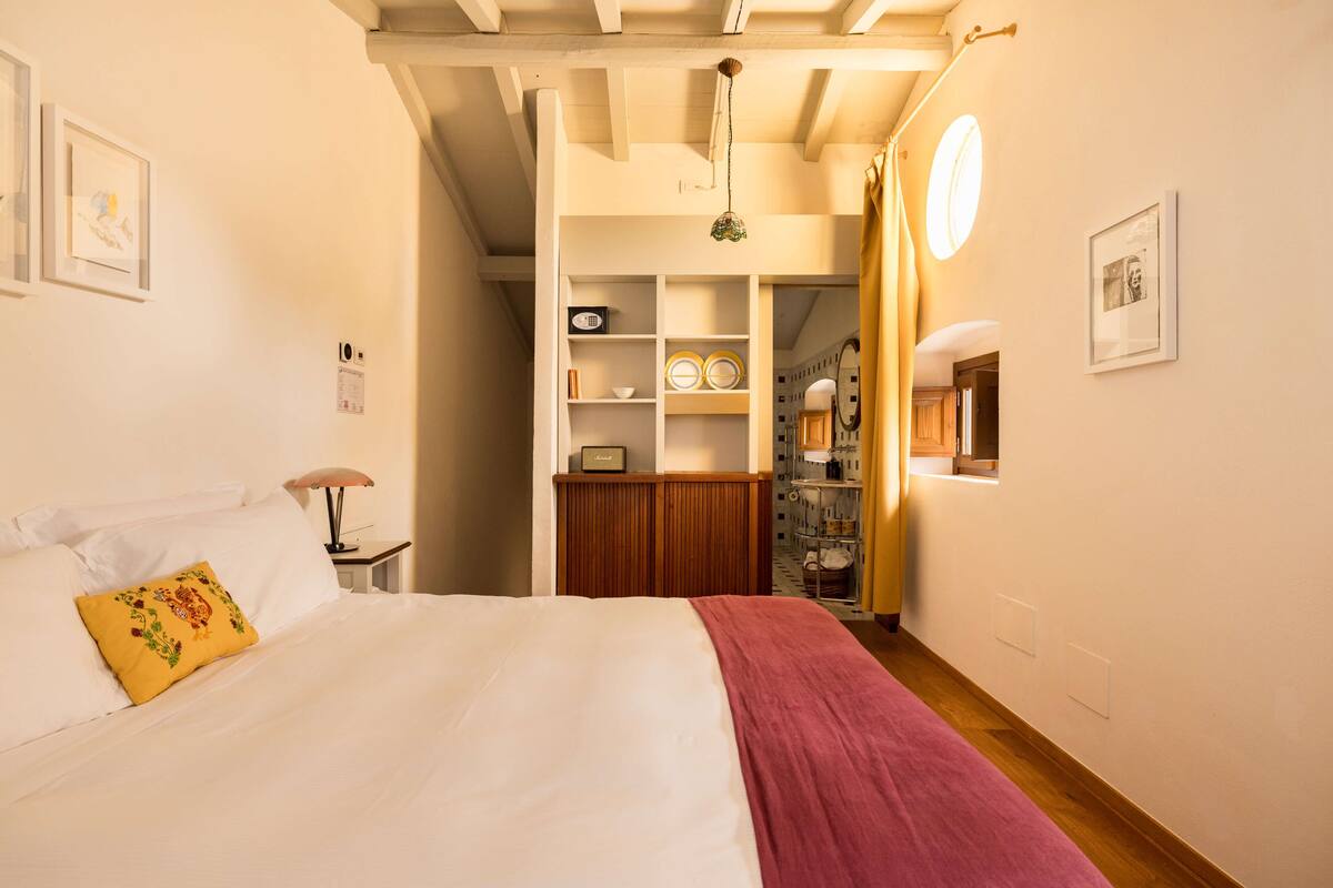 Un dormitorio con cama, mesita de noche, lámpara, estante con adornos y una puerta que da al baño.