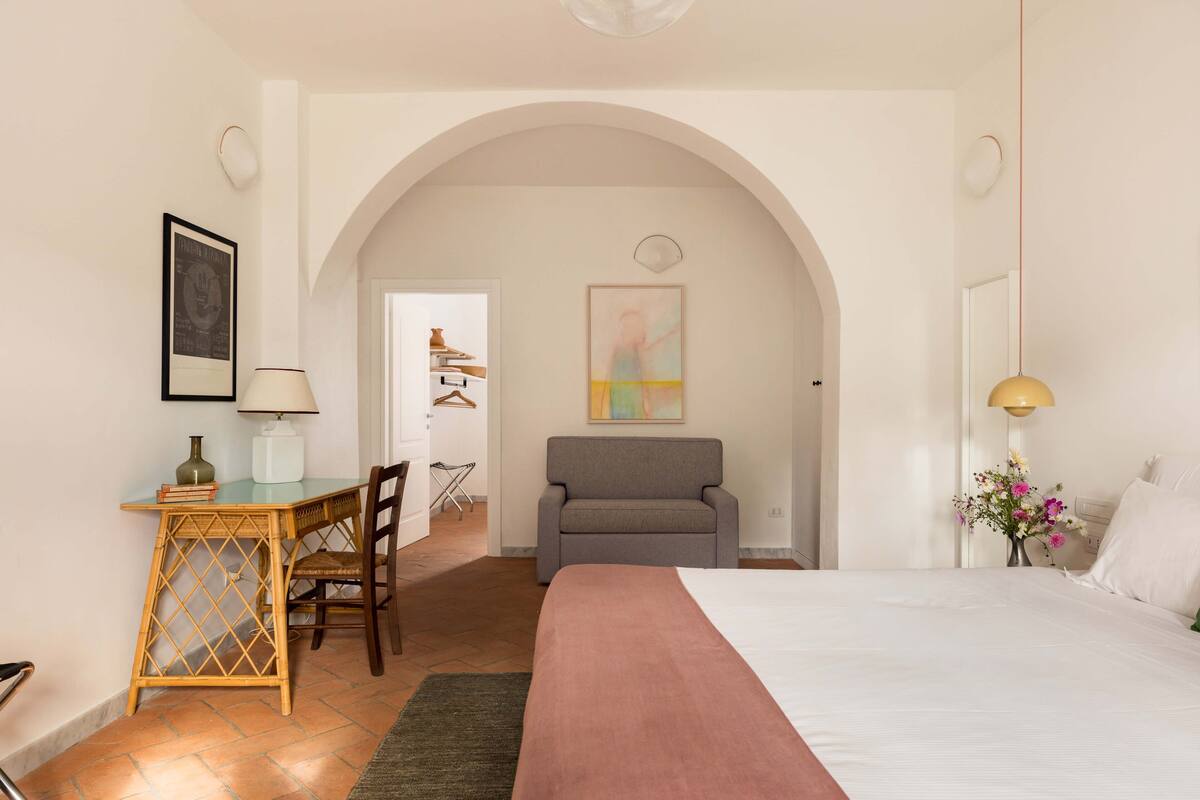 Un dormitorio con cama, escritorio con lámpara, silla y una pequeña mesa con libros.