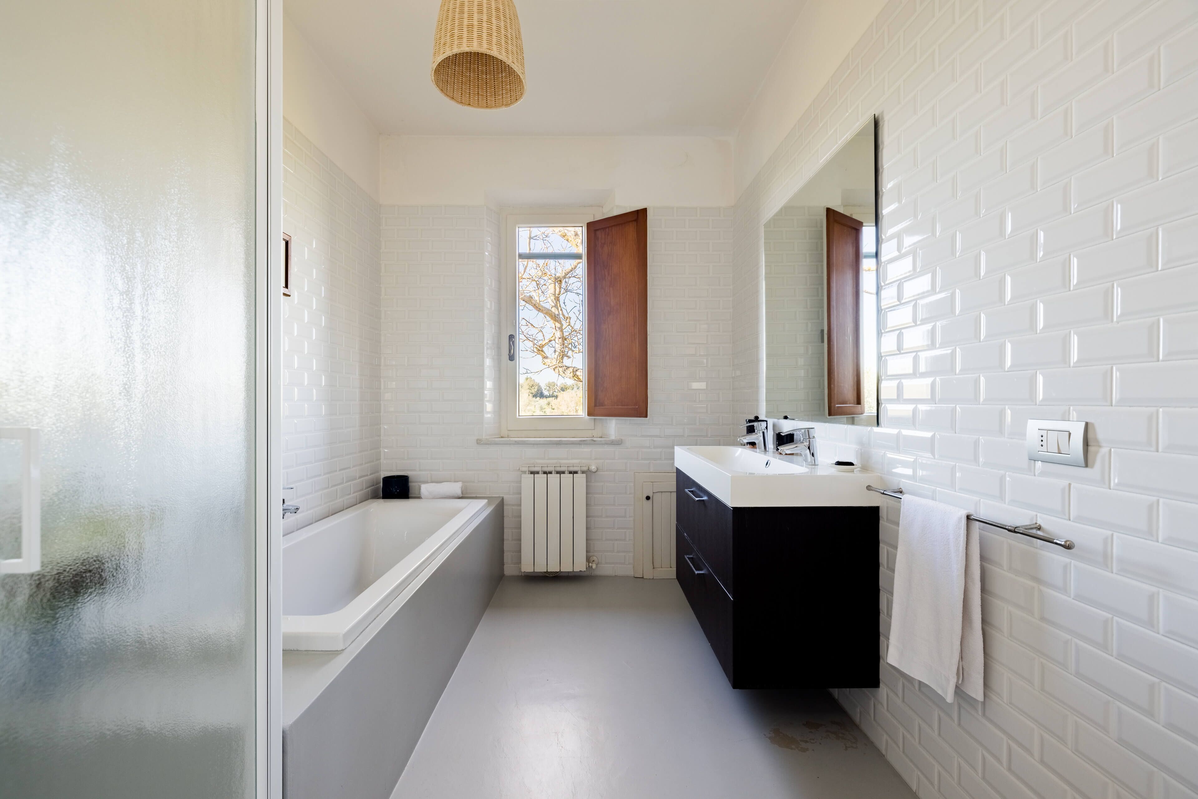 Ein modernes Badezimmer mit weißen Fliesen, einer Badewanne, einem Waschbecken mit Spiegel und einem Fenster mit Blick auf Bäume.