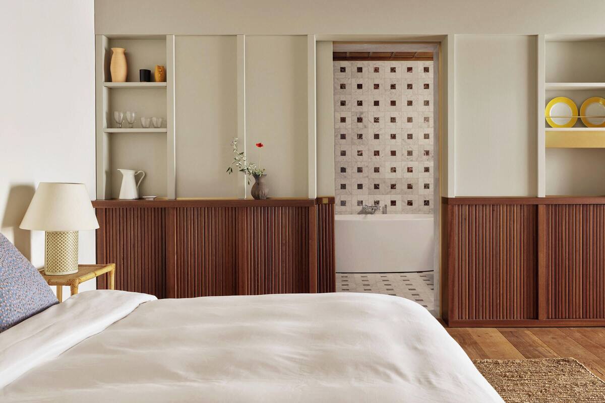 Habitación de hotel con cama, mesita de noche con lámpara, aparador de madera, estante con cristalería y baño con zona de ducha azulejada.