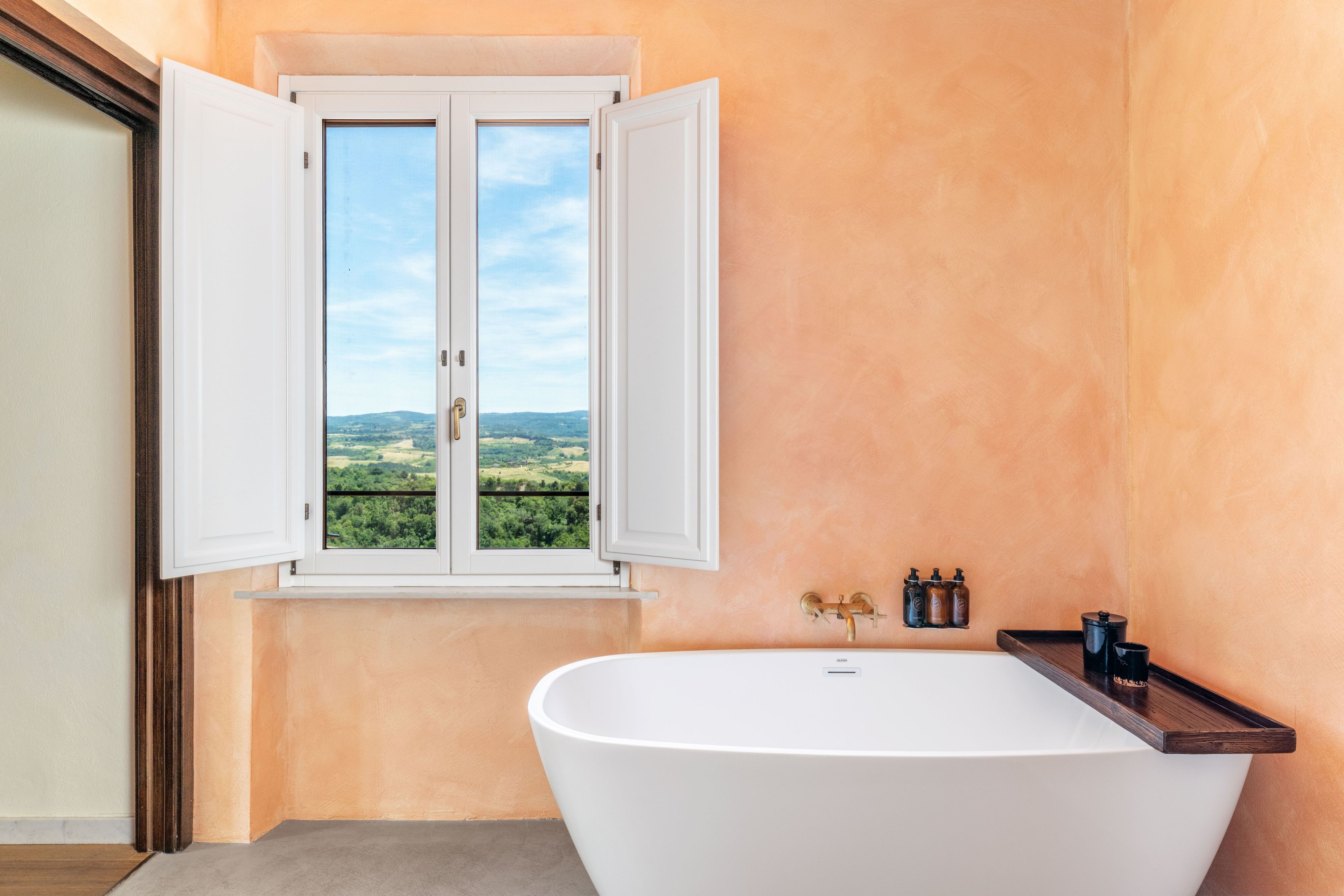 A bathroom with a bathtub, a window with open shutters, and a view of a green landscape.