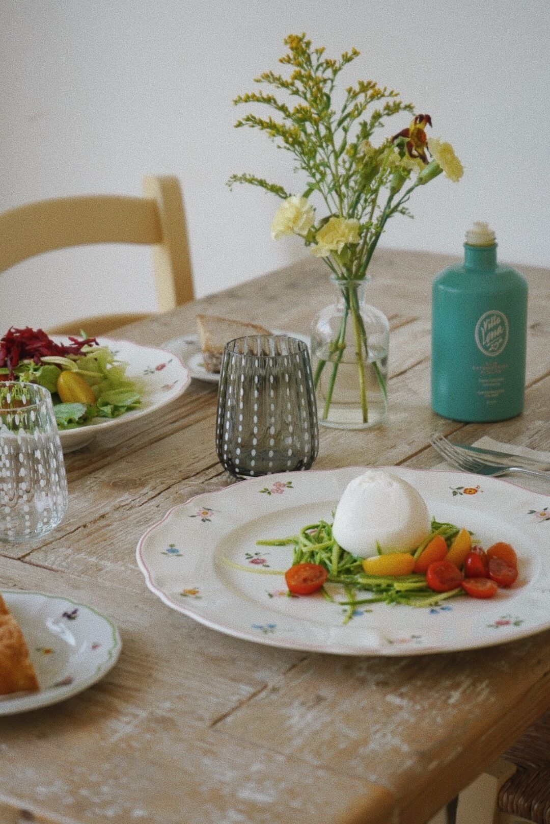 Una mesa de comedor con platos de comida, un jarrón con flores y una botella.