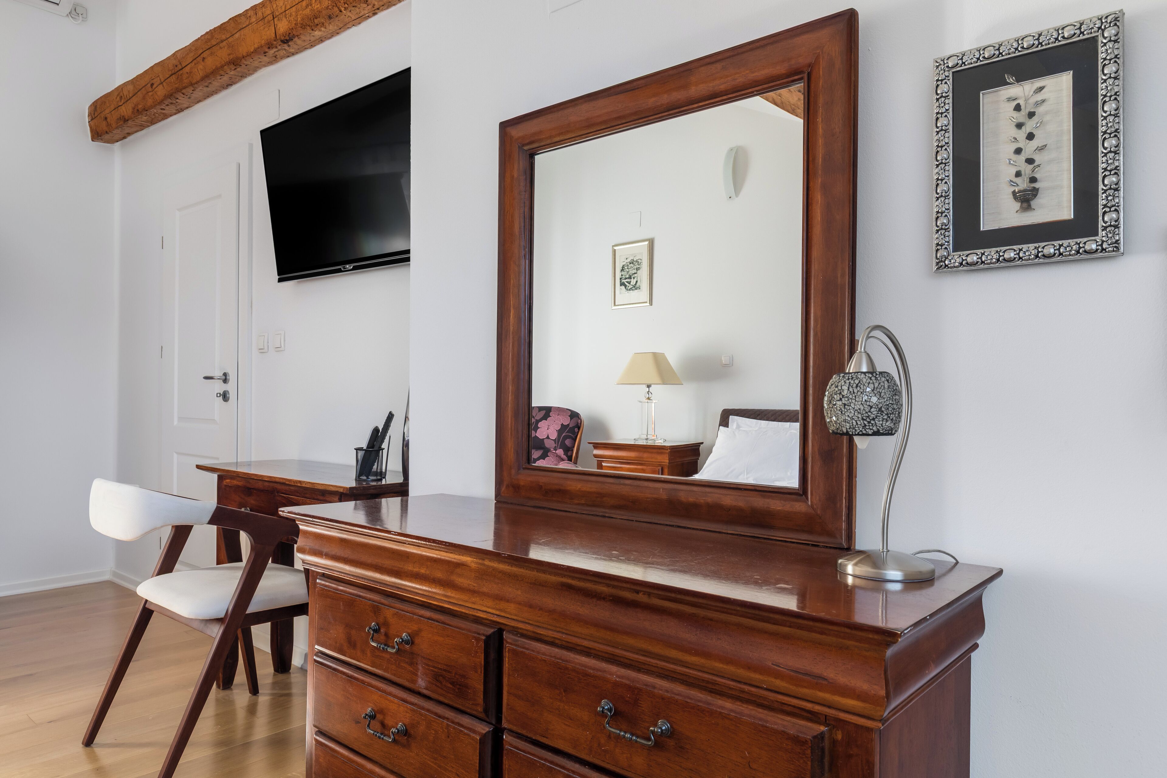A wooden dresser with drawers, a mirror, a framed picture, a lamp, and a television on the wall.