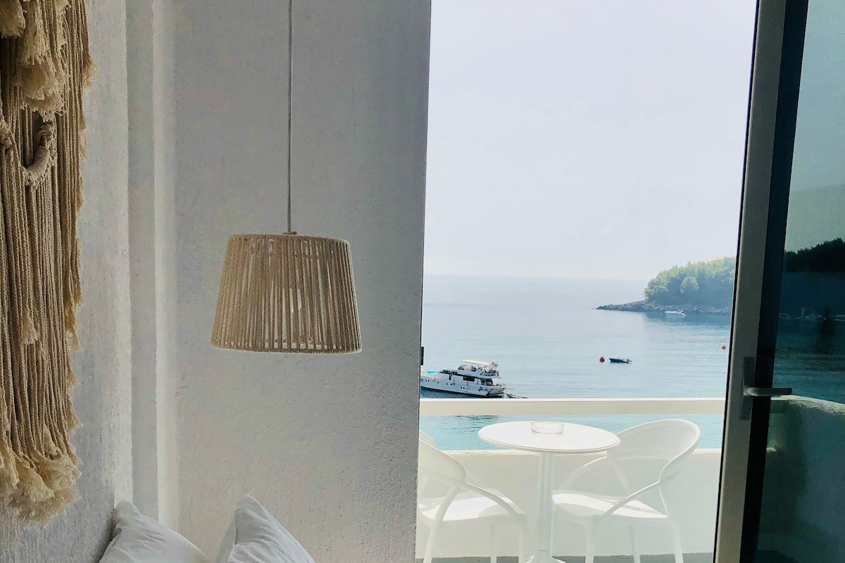 Ein Schlafzimmer mit großem Fenster, das auf einen Balkon mit Tisch und Stühlen sowie Blick auf das Meer hinausblickt. Eine Hängelampe ist ebenfalls zu sehen.
