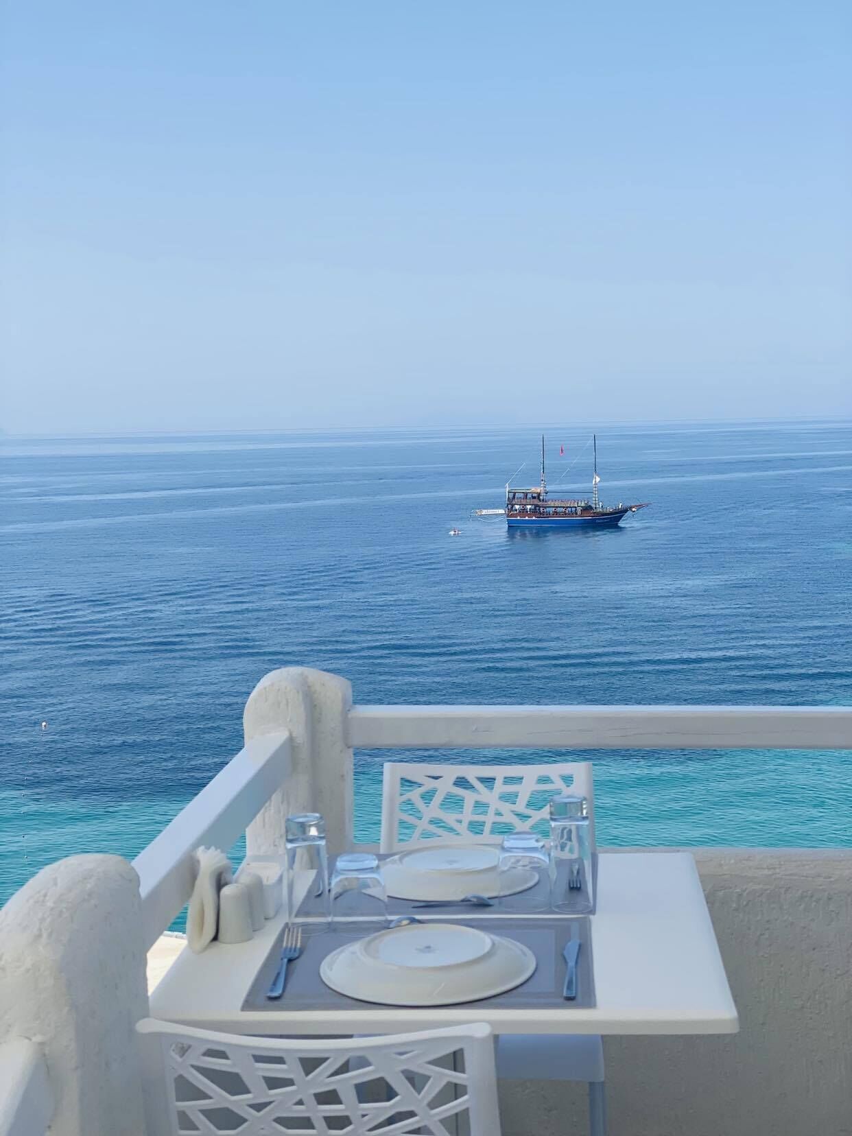 A table set with plates, cutlery, and glasses on a balcony overlooking a calm sea with a boat in the distance.