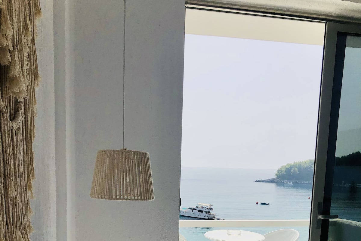 Ein Schlafzimmer mit einem großen Bett, weißen Kissen, einer Wanddeko aus Makramee, einer Hängeleuchte und Meerblick durch eine Schiebetür.