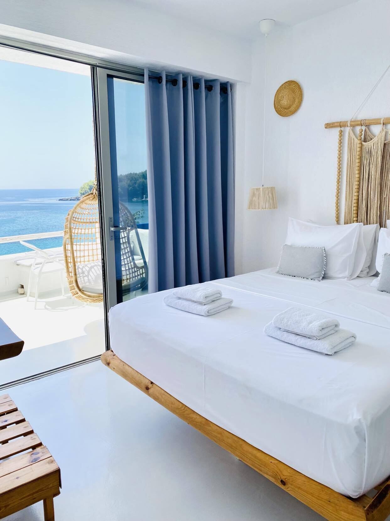 A hotel room with a large bed, a view of the sea, and a wicker chair outside the window.