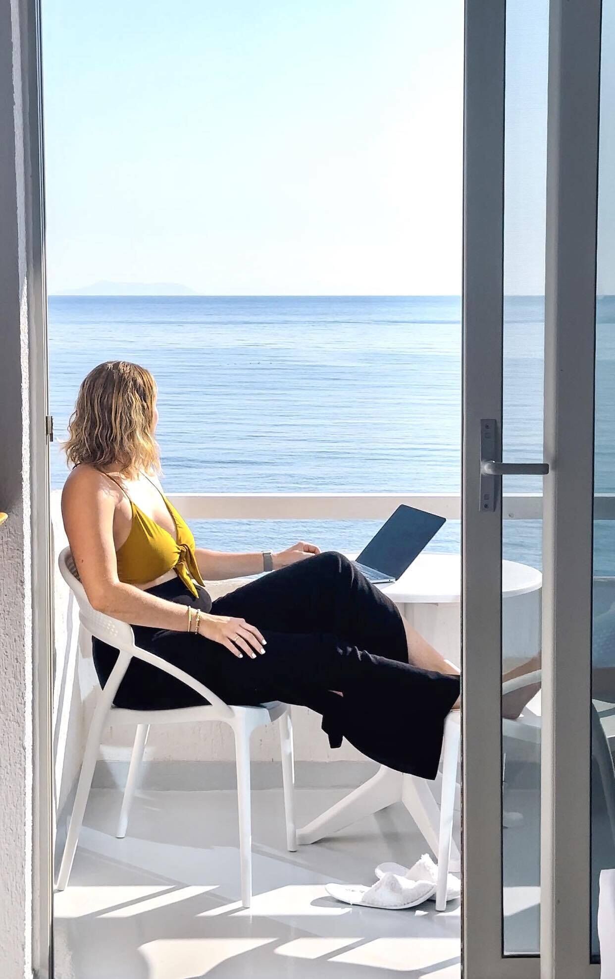 A woman sitting on a white chair with a laptop on a balcony overlooking the sea.