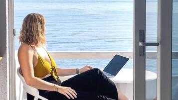 A woman sitting on a white chair with a laptop on a balcony overlooking the sea.