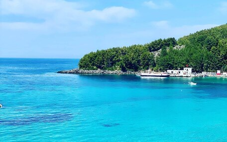 A coastal view with clear turquoise waters, a rocky shoreline, and a small building with a red roof on a hill.