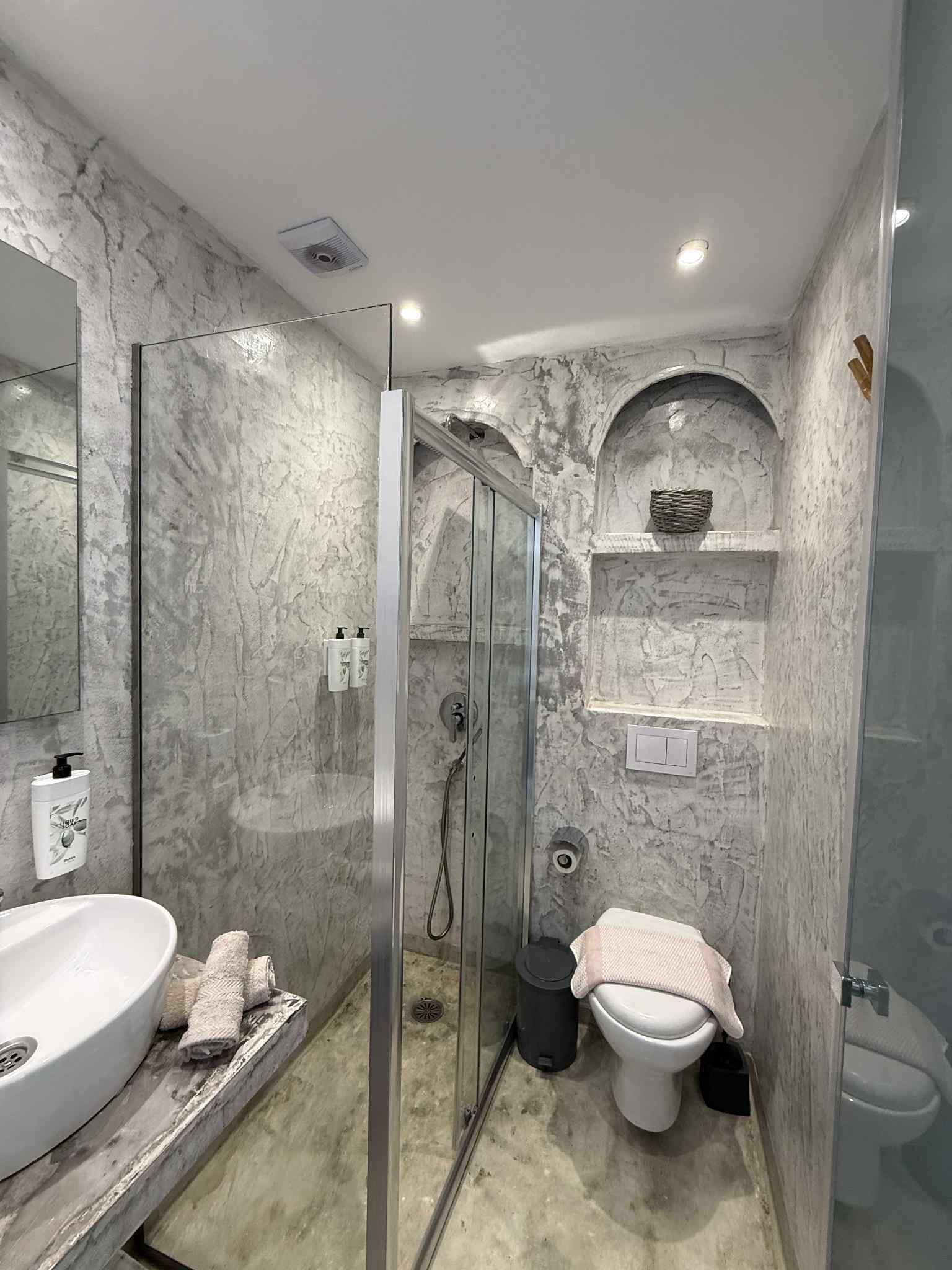 A bathroom with a marble wall, a glass-enclosed shower, a white sink, and a toilet.
