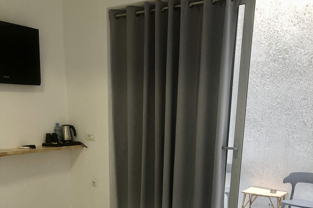 Ein Hotelzimmer mit einem Bett, einem kleinen Tisch, einem Sessel, einem an der Wand montierten Fernseher und einem Fenster mit Vorhängen.