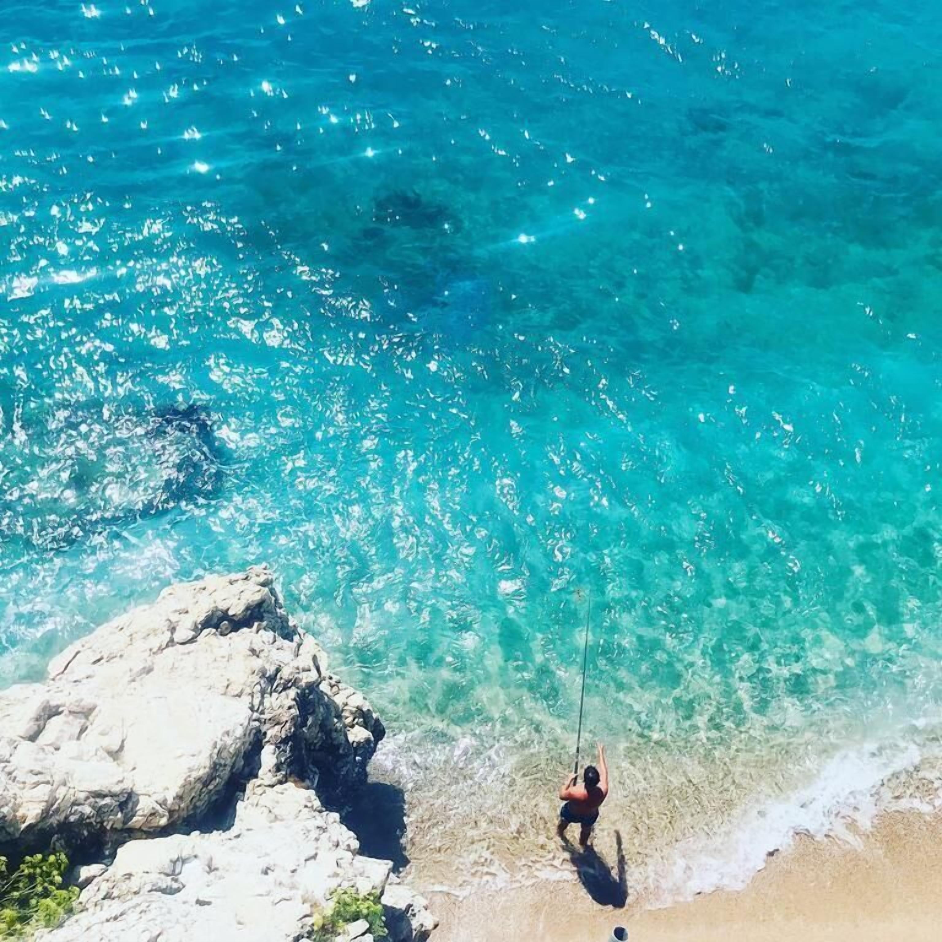A person fishing at a beach with clear turquoise water and rocky formations.