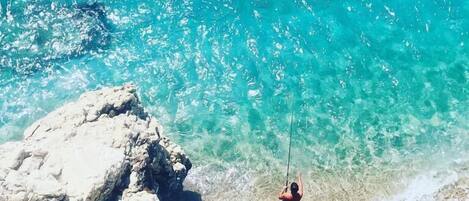 A person fishing at a beach with clear turquoise water and rocky formations.