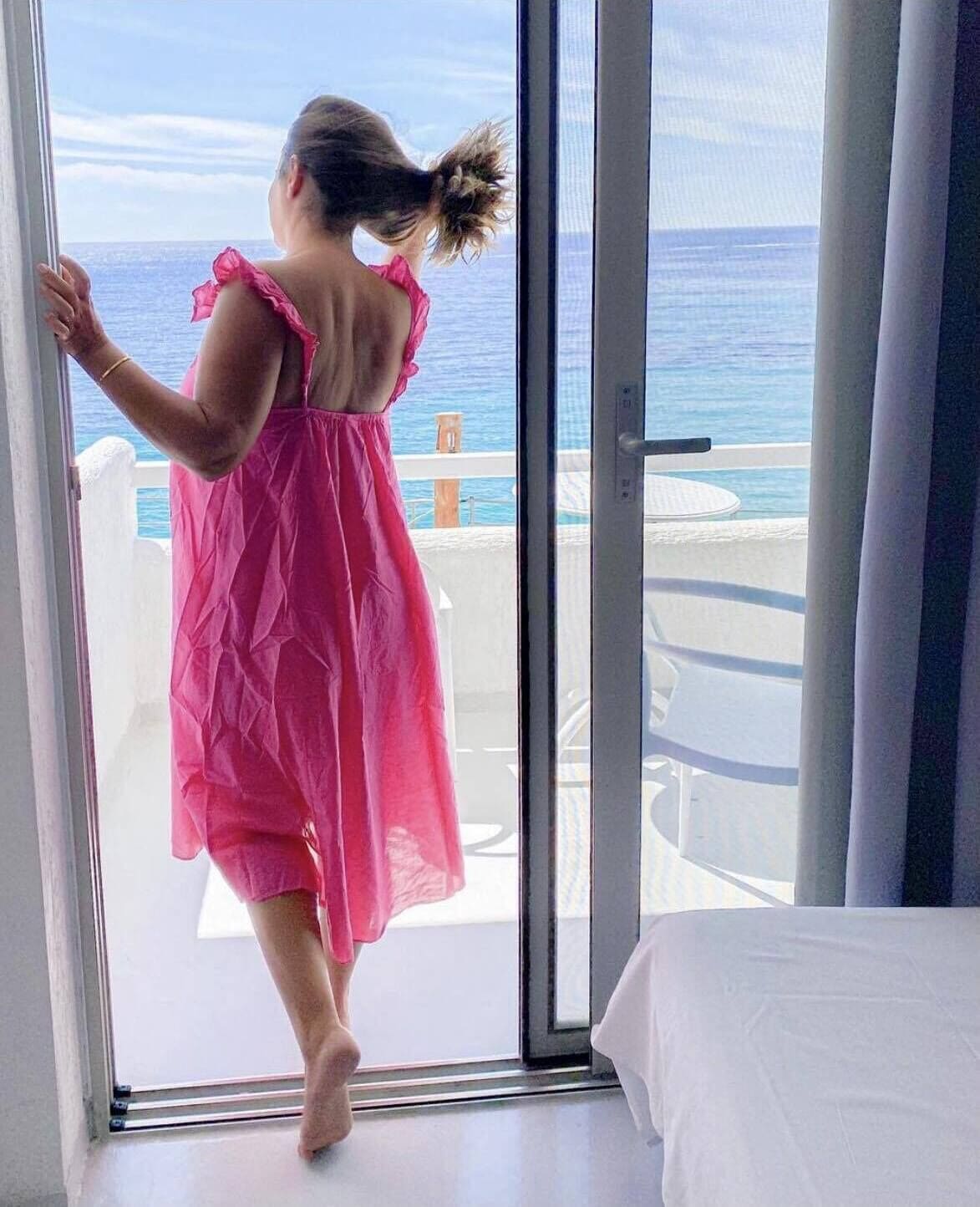 A person in a pink dress standing at a sliding glass door with a view of the ocean.