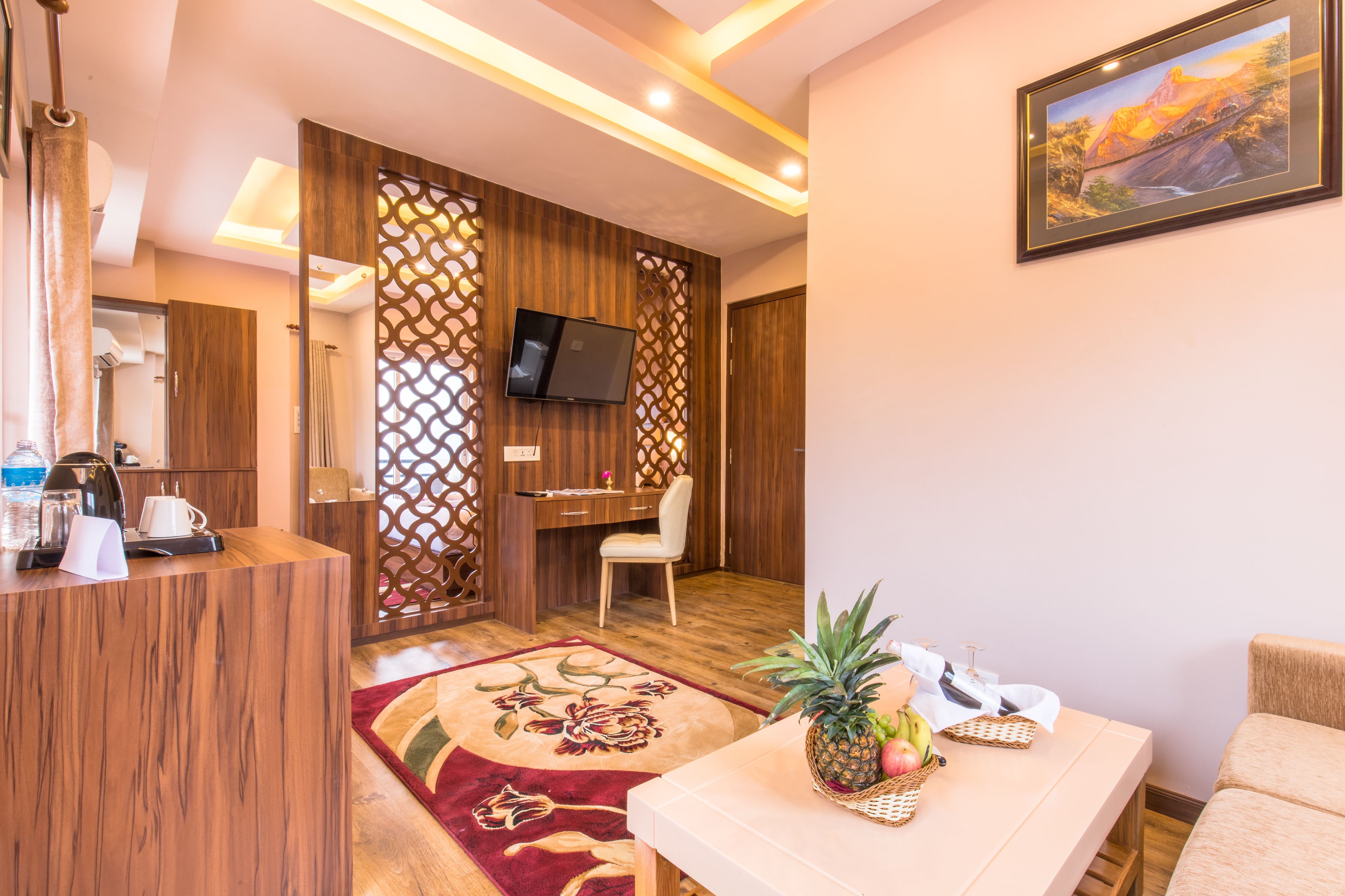 Habitación de hotel con una recepción de madera, un televisor montado en un panel de madera, un escritorio con una silla, una pequeña mesa con una piña y frutas, y un cuadro en la pared.