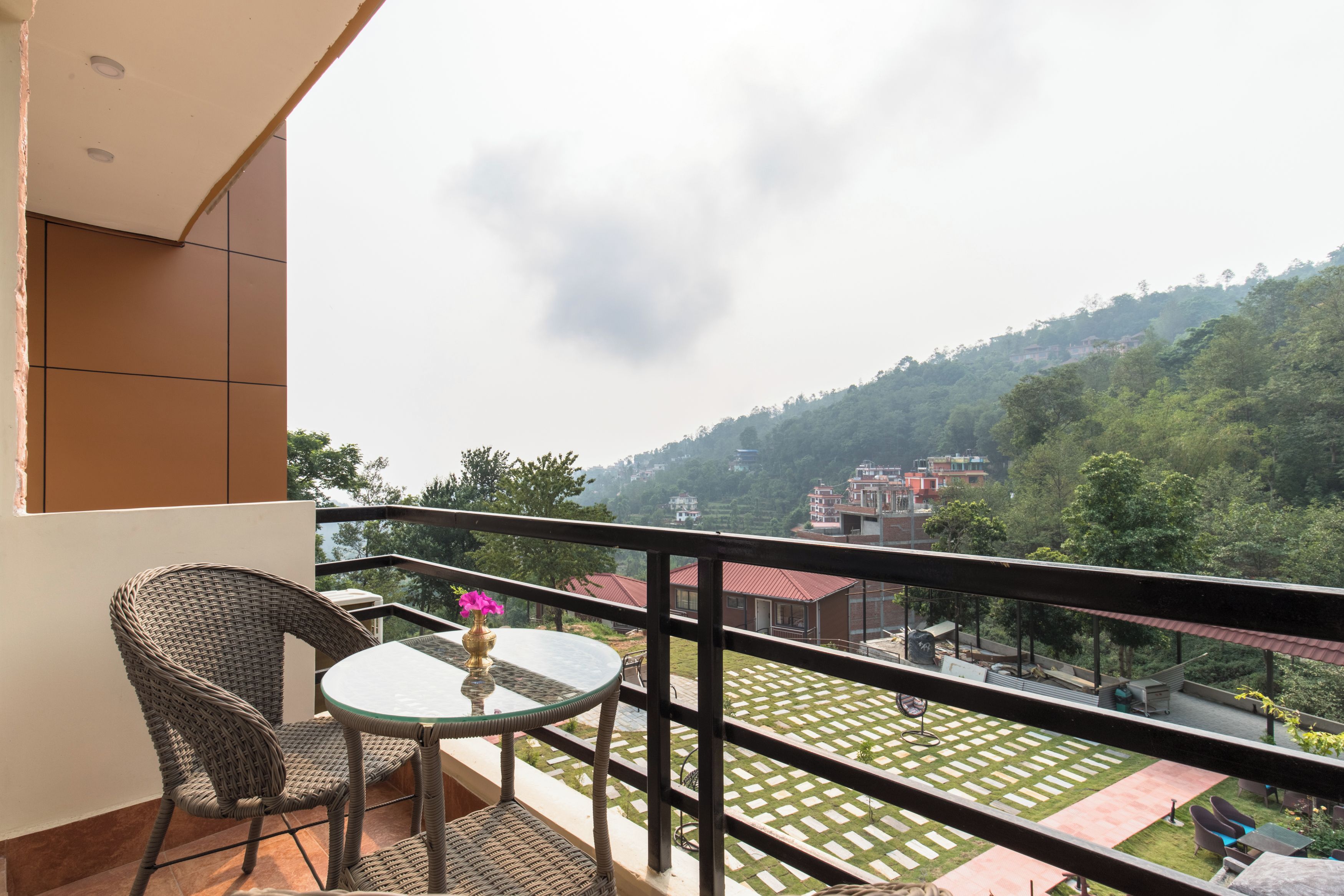 A balcony with wicker furniture, a glass table, and a view of a green landscape with buildings.