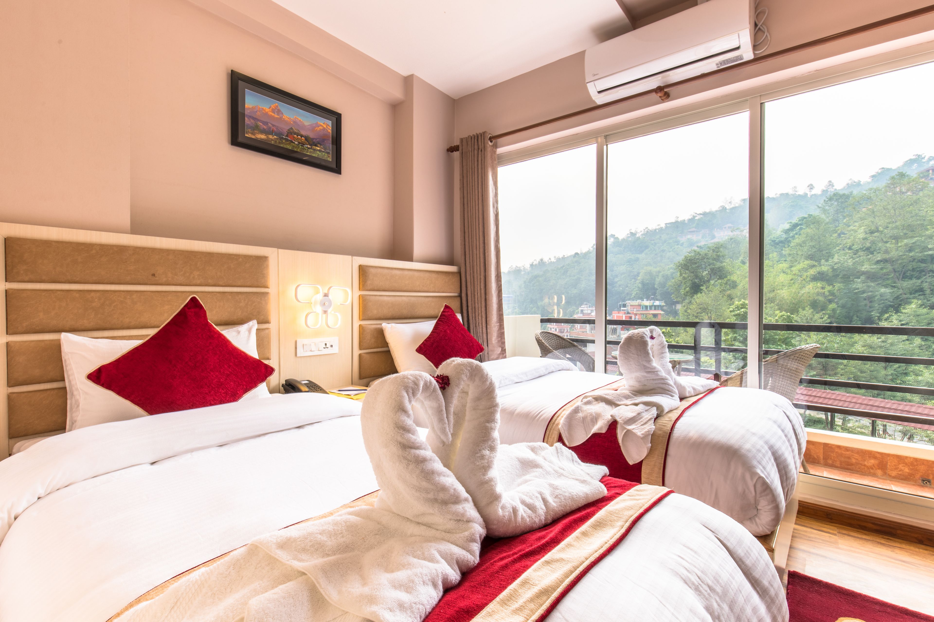Una habitación de hotel con dos camas, cada una con ropa de cama blanca y almohadas rojas, un ventanal con vistas a una ladera boscosa y un equipo de aire acondicionado en la pared.