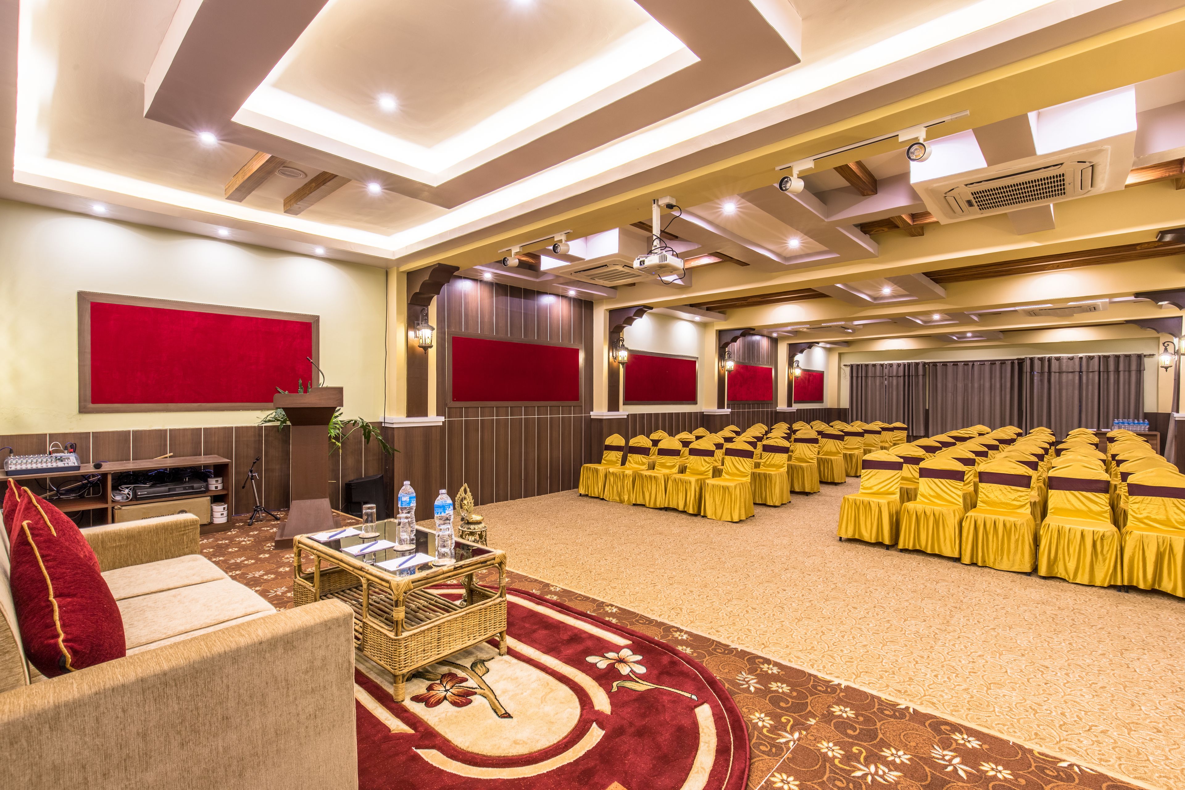 Un salón de conferencias con filas de sillas de color amarillo, un escenario con fondo rojo y una zona de asientos con alfombra roja y mobiliario de mimbre.