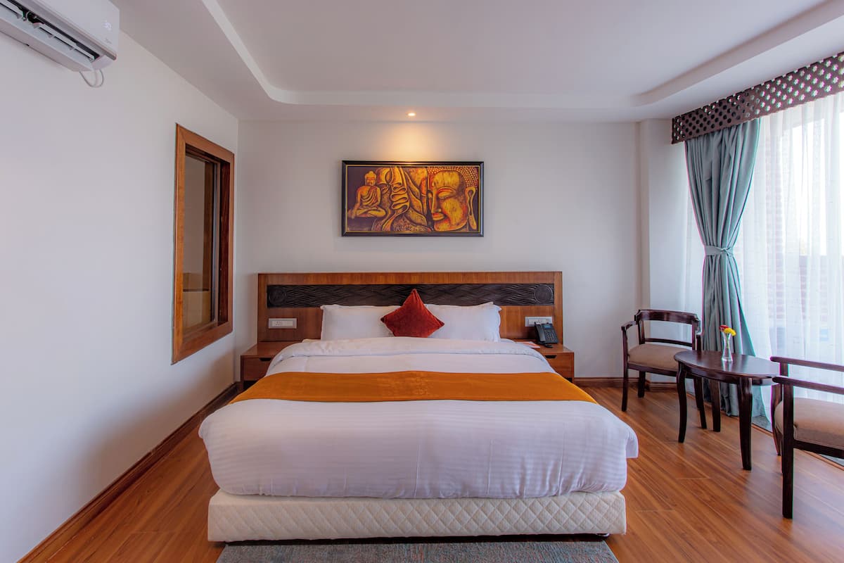 Quarto de hotel com uma cama grande, cabeceira de madeira, pintura sobre a cama, escrivaninha com cadeira e janela com cortinas.