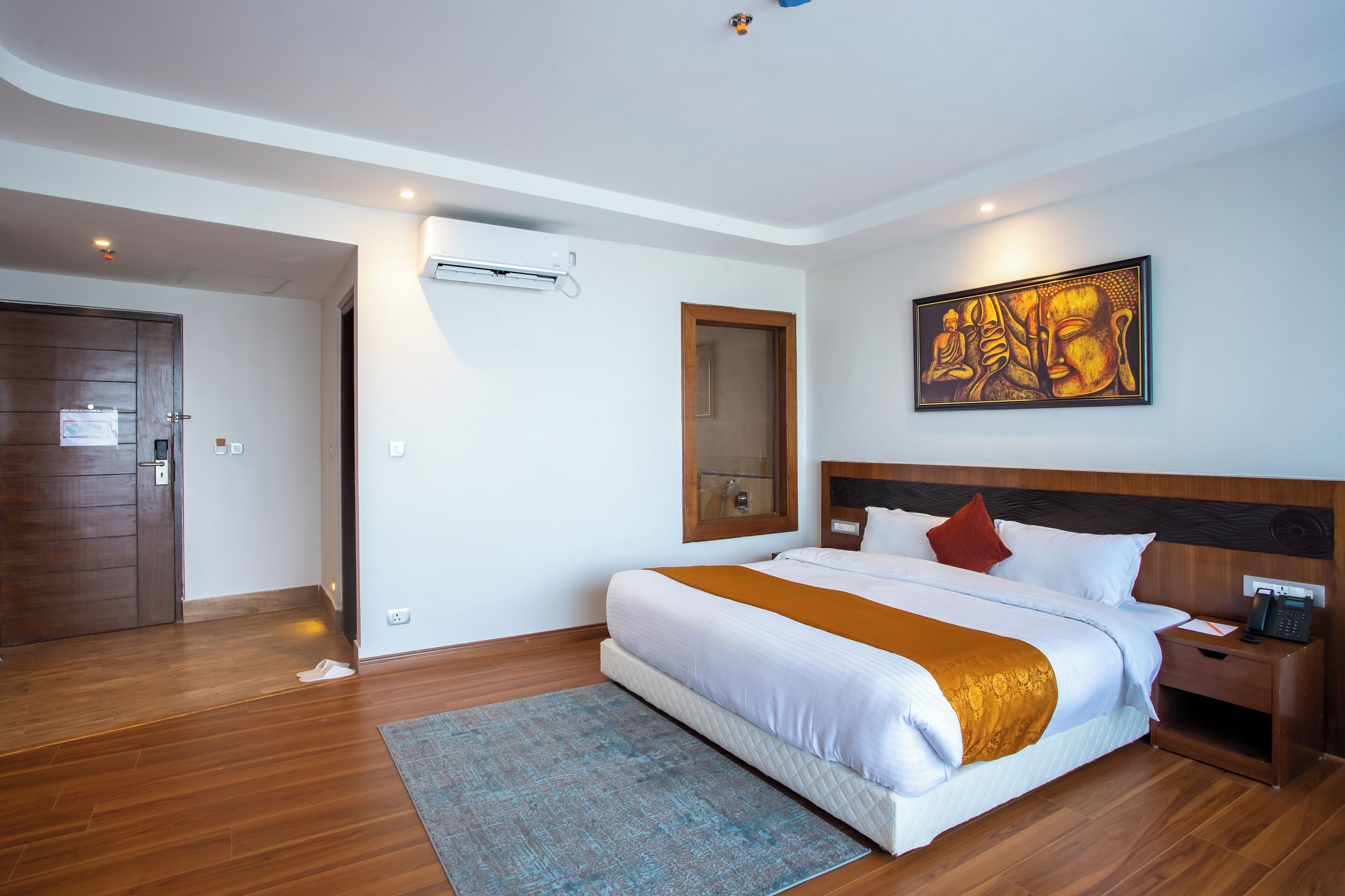 A hotel room with a large bed, wooden flooring, a wall-mounted air conditioner, a painting, and a nightstand with a phone.