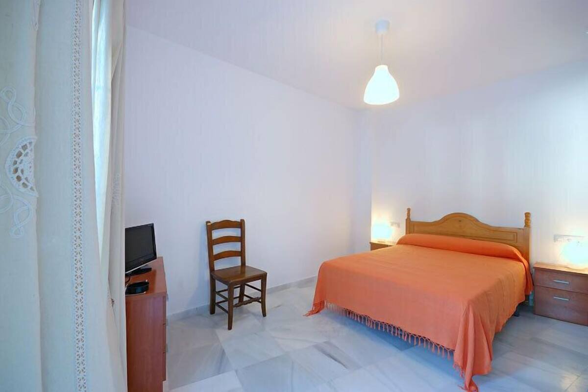 A bedroom with a bed, a chair, a desk with a TV, and a lamp.