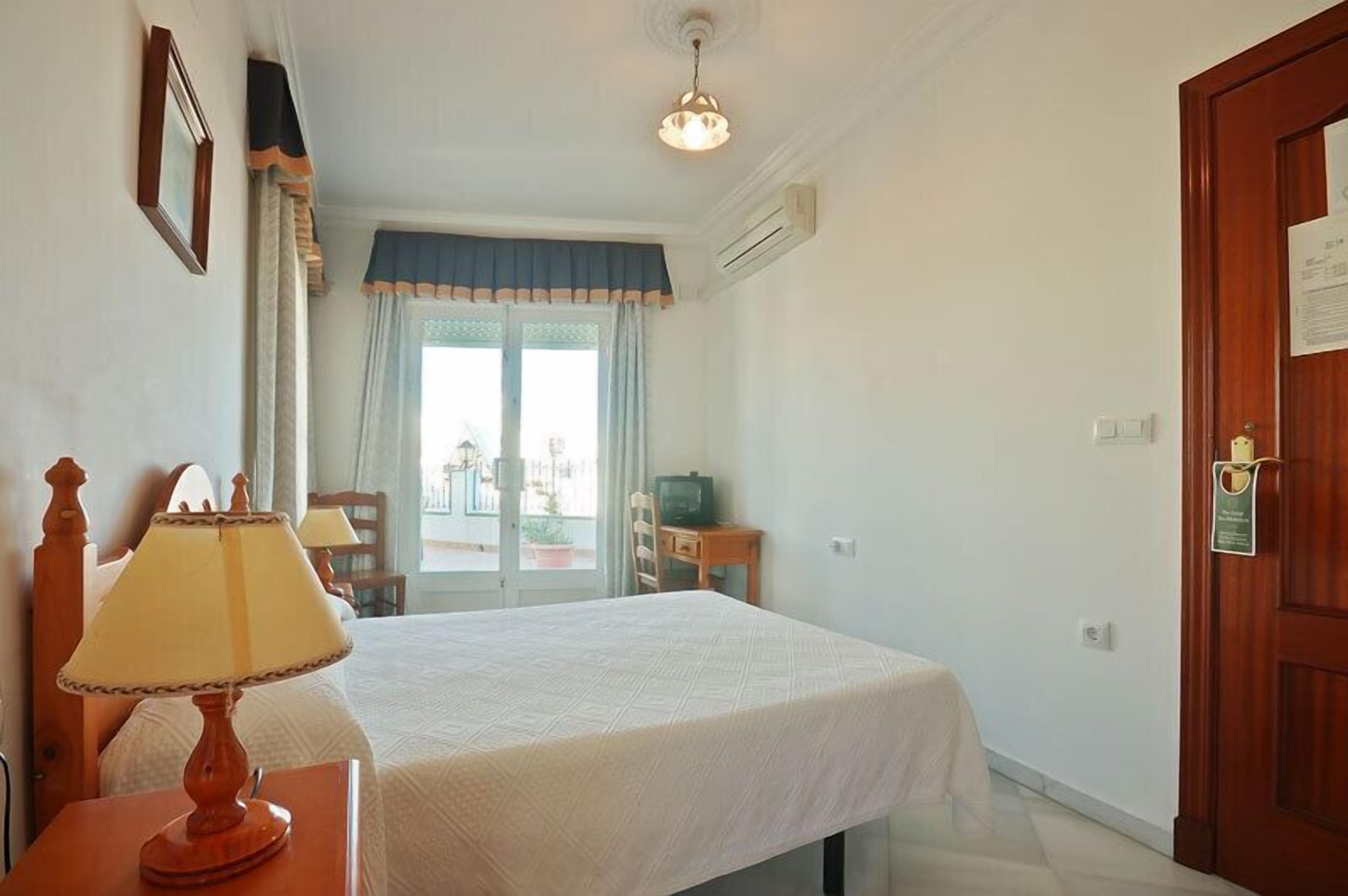A hotel room with a bed, a desk with a lamp, a television, and a view of the outside.